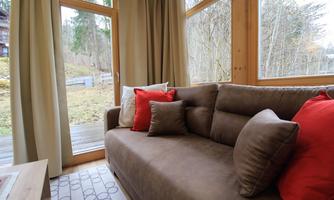 A cozy sofa with red and brown cushions is in a bright room. Large windows offer a view of the nature outside.