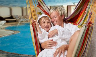 A mother and her daughter relax in a hammock by the pool. Both are wearing bathrobes and are happily smiling at the camera.