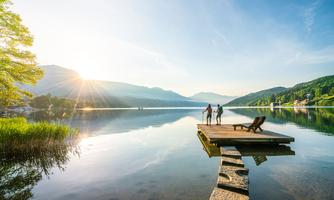 A picturesque lake with a wooden pier and gentle mountains in the background. The sun rises and illuminates the calm water surface.