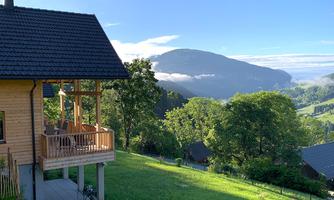 A cozy wooden house with a balcony in a green landscape. In the background, gentle hills and a clear blue sky can be seen.