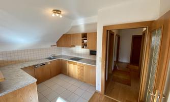 A modern kitchen with wooden cabinets and light flooring. The room is well-lit and offers a view of the hallway.