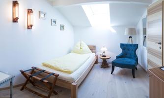 A cozy bedroom with a single bed, a blue armchair, and a small table. The walls are light, and the skylight provides plenty of daylight.