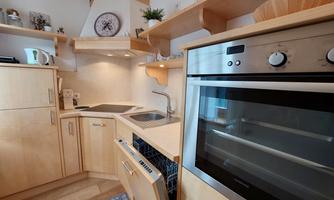 A modern kitchen with wooden furniture and stainless steel appliances. The cabinets and floor are light, and there is a small cooking area.
