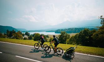 Two people are riding bicycles on a road next to a clear lake. In the background, mountains and a green landscape can be seen.