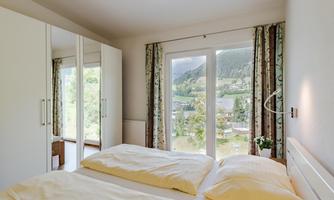 A cozy bedroom with a large window and a view of the mountains. The decor is bright and modern, featuring a double bed and a wardrobe.