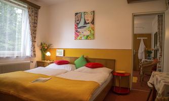 A cozy room with two beds and colorful cushions. Pictures hang on the walls and there is a small table decorated with a plant.