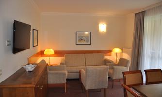 A cozy living room with a sofa, two armchairs, and a dining table. The walls are bright and there is a TV setup as well as ambient lighting.