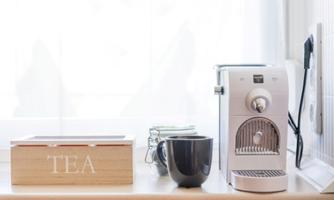A modern kitchen corner with a coffee machine and a tea box. A black cup sits next to the machine on the countertop.