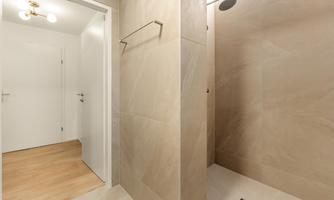 A modern bathroom with a shower made of light tiles. The entrance door is visible and the floor is made of wood.