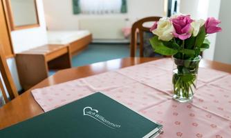 An inviting table with a green welcome folder and a vase full of pink and white roses. In the background, a simply furnished room is visible.
