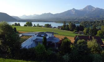 A picturesque landscape with a clear lake and green meadows. In the background are mountains and a quiet residential area.