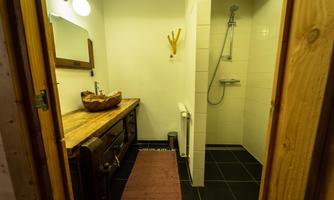 A modern bathroom with a wooden dresser and a large mirror. On the right, a shower is installed, and the floor is equipped with dark tiles.