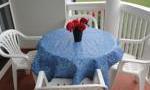 A round table with a blue tablecloth is on a balcony. On the table, there is a vase with red flowers.
