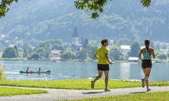 Two women are jogging by the shore of a clear lake. In the background, a church and some houses can be seen in a beautiful landscape.
