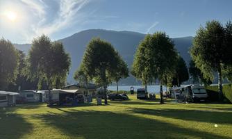A campsite with green meadows and trees. In the background, mountains and a calm lake can be seen.