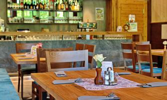 A cozy restaurant with wooden tables and chairs. In the background, there is a well-stocked bar with drinks.