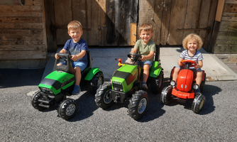 Three children are sitting on small tractors in front of a wooden barn. The sun is shining and the atmosphere is cheerful and playful.