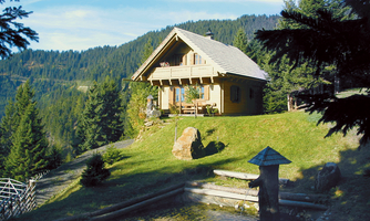 A beautiful wooden house surrounded by green mountains and trees. In the foreground, there is a small pond.