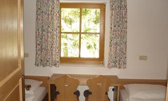 A simple room with two single beds and a table between them. A large window with curtains lets in plenty of daylight.