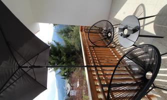 A balcony with two black chairs and a table. A parasol provides shade, while trees are visible in the background.