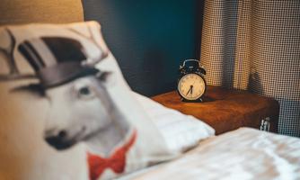 A cozy bedroom with a cushion that shows a stag in a hat. Next to the bed, there is a stylish alarm clock on a wooden table.