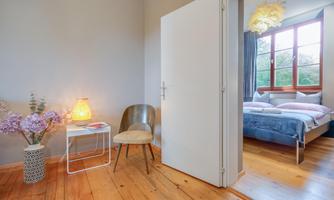An inviting hall with a chair and a lamp. In the background, you can see a cozy bedroom with a large window.