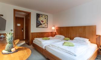 A cozy hotel room with two single beds and modern wooden interiors. A piece of art hangs on the wall, and fresh fruit is on the table.