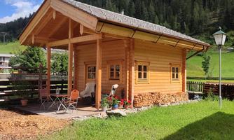 A cozy wooden cabin stands in a green setting. In the background, forested hills and a blue sky can be seen.