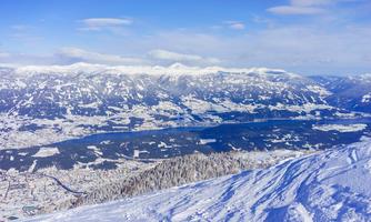 An impressive winter landscape with snow-covered mountains and a clear blue sky. In the foreground, rolling snowfields can be seen, and in the background, a clear lake stretches out.