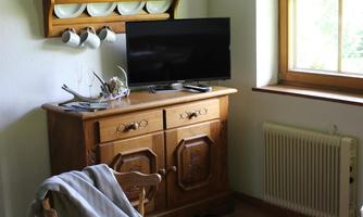 A cozy room with a wooden cabinet and a television. The window provides plenty of light, and in the background, there are plates on a shelf.