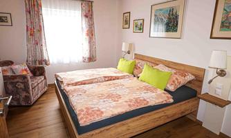 A cozy bedroom with a large bed and colorful pillows. The walls are decorated with pictures, and an armchair is in the corner.