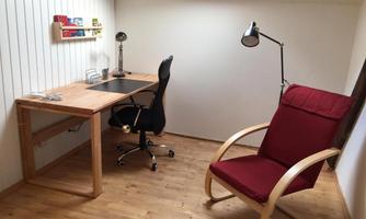 A cozy study with a wooden desk and a swivel chair. Next to the desk is a red armchair, and a lamp provides light.