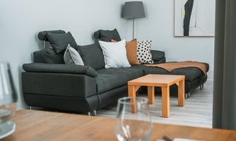 A modern living room with a gray sofa and colorful cushions. A small wooden table stands in front of the sofa, and a floor lamp provides cozy light.