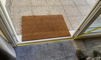 A doormat in front of an open door. The floor is made of gray tiles and there is a rug indoors.