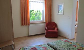 A bright room with a double bed and a red armchair. The window lets in plenty of daylight and offers a view of outdoor plants.