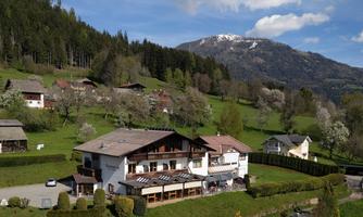 A well-maintained building in a green landscape with mountains in the background. There are numerous trees and blooming plants in the vicinity.