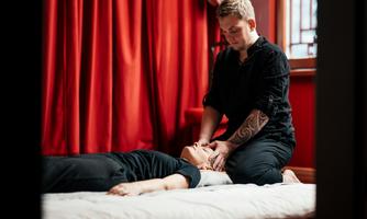 A masseur performs a relaxing massage on a lying person. The room is decorated with red curtains, creating a calming atmosphere.
