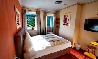 A cozy room with a large bed and a window front. The walls are in warm tones, and a TV as well as decorative pictures are present.