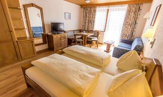 A cozy hotel room with two beds, a television, and a small dining table. Large windows provide plenty of daylight, and the decor is simple and inviting.