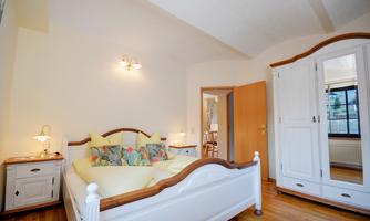 A bright bedroom with a white bed and colorful pillows. The room has a wooden floor and a wardrobe.