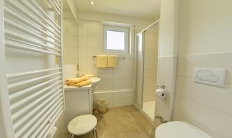 A modern bathroom with light tiles and plenty of natural light. There is a shower, a sink, and towels on a shelf.