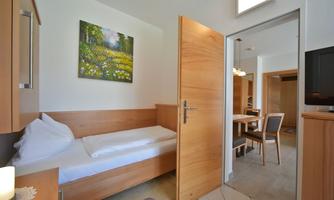 A cozy room with a single bed and a small table.  
On the wall hangs a picture of a blooming meadow, and the door leads to another room.