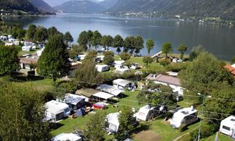 A picturesque campsite by the shore of a lake, surrounded by trees and mountains. The caravans and tents are neatly arranged and invite relaxation in nature.