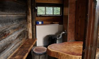 A rustic interior view of a small room with wooden paneling. There is a round wooden table, seating, and a window looking outside.