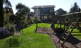 A beautiful garden with a playground and colorful flowers. In the background, there is a house under a clear blue sky.