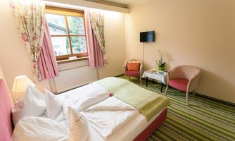 A cozy hotel room with a double bed, a seating area, and colorful curtains. The room features a television and a pleasant green carpet.