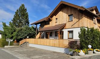 A modern wooden house with a bright facade and a spacious balcony. Surrounded by green trees and flower beds.