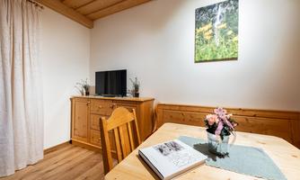 A cozy living room with a wooden table, a television, and a flower vase. A painting of a mountain landscape hangs on the wall.