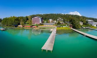 A picturesque lake with clear, blue water and a wooden jetty. In the background, green trees and beautiful buildings can be seen.