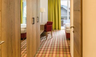 A bright hallway with wooden paneling and a colorful rug. On the left side are doors, and at the end of the hallway stands a red chair next to a window with green curtains.